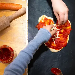 Kinderhände streuen geriebenen Käse über ausgerollten Teig mit Tomatensauce