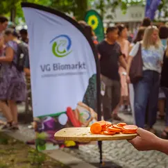 eine Hand hält in Brett mit geschnittenen Möhren darauf, im Hintergrund eine Bowflag mit dem Logo der Verbrauchergemeinschaft.