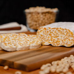 frischer aufgeschnittener Tempeh auf einem Brett, im Hintergrund ein Glas mit Sojabohnen