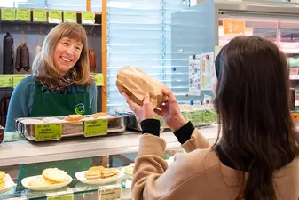 Kundin an der Theke hält eine gefüllte Tüte in der Hand, eine Mitarbeiterin lächelt sie an