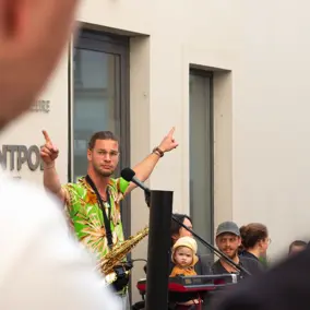 Musiker steht an einem Mikrofon und hebt die Arme, daneben zuhörende Menschen