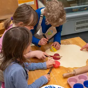 Drei Kinder bereiten beim Kochworkshop ausgerollten Pizzateig auf einem Tisch vor