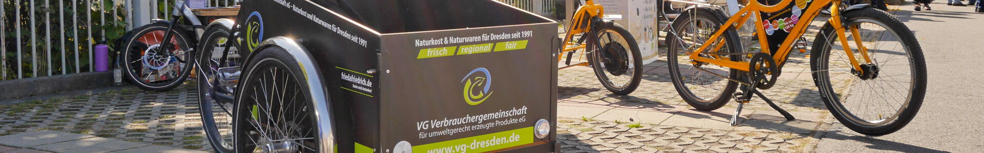 Fahrräder und ein Lastenrad im Vordergrund, bedruckt mit dem farbigen Schriftzug "VG Verbrauchergemeinschaft für umweltgerecht erzeugte Produkte eG"