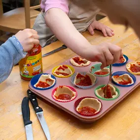 Drei Kinder befüllen ein Muffinblech mit Pizzateig und Tomatensauce