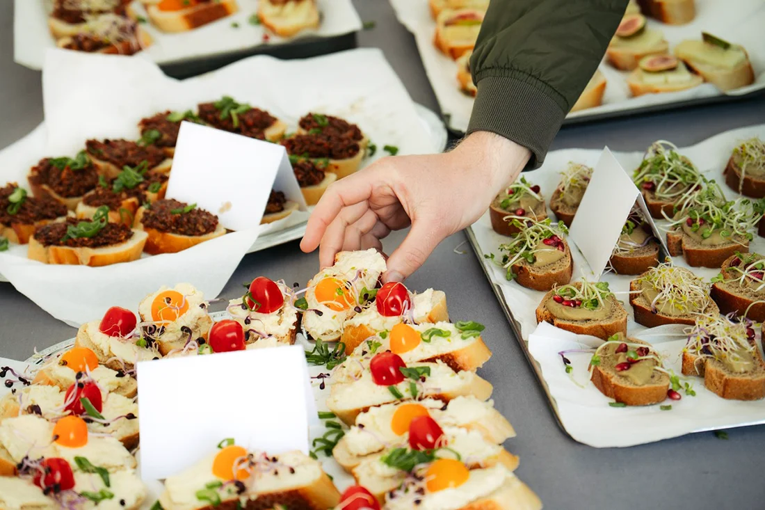 Snacks und Schnittchen auf drei Tabletts, eine Hand greift nach einem der Schnittchen.