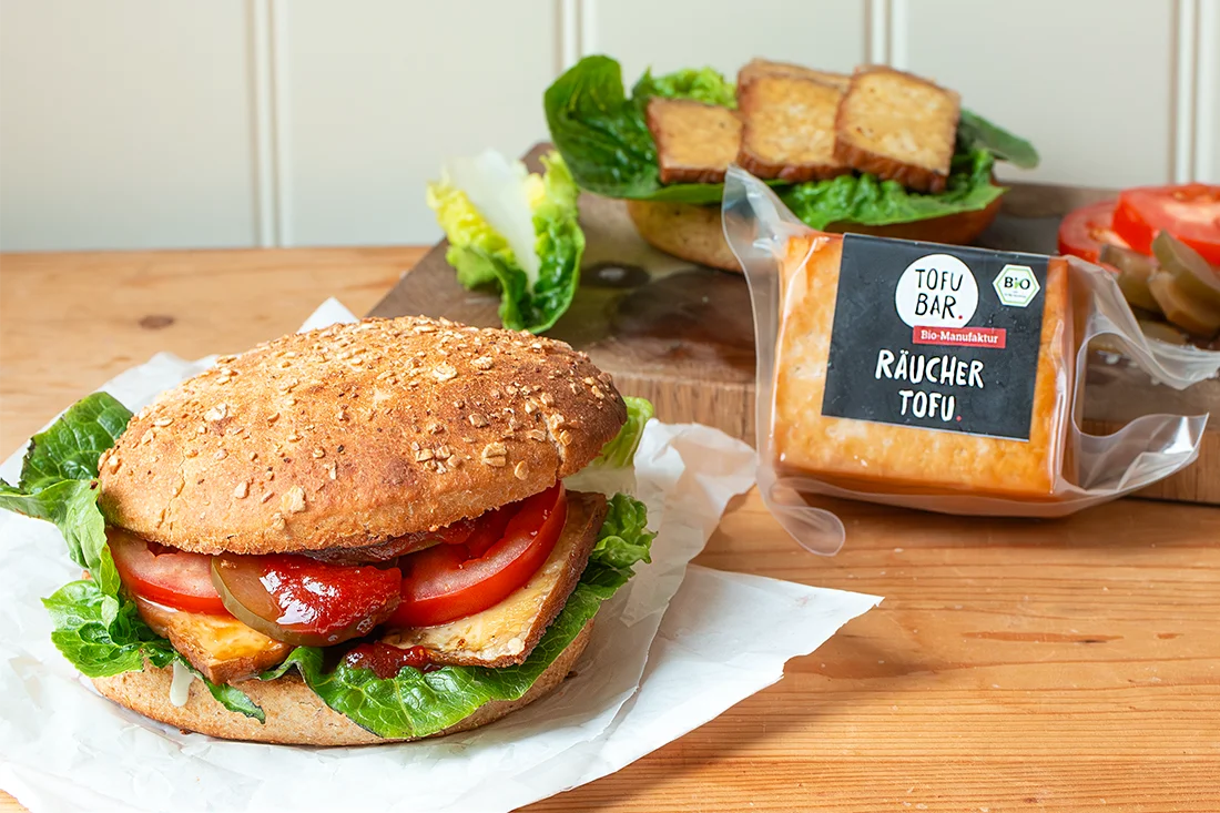 Burger mit Räuchertofu, Tomaten und Salat belegt, im Hintergrund Räuchertofu von Tofubar