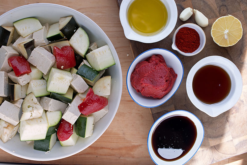 Geschnittene Zucchini, Aubergine, Paprika und Tofu in einer Schüssel, daneben Tomatenmark, Öl, Sojasauce, Sirup, Gewürze, Knoblauch und Zitrone in Schälchen