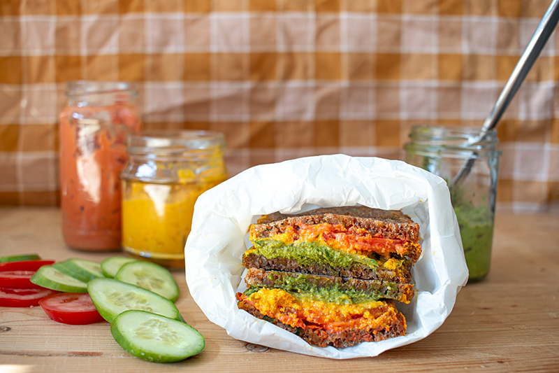 Pausenbrot mit bunten Aufstrichen, Gurkenscheiben, Tomatenscheiben und drei Gläser mit Aufstrichen auf einem Holzbrett
