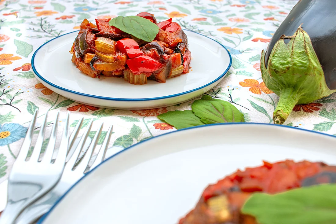 Auberginen Caponata auf einem Teller, daneben eine Aubergine und Besteck