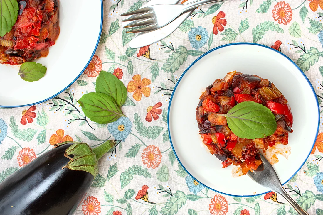 Caponata auf einem Teller mit einer Gabel, daneben eine Aubergine