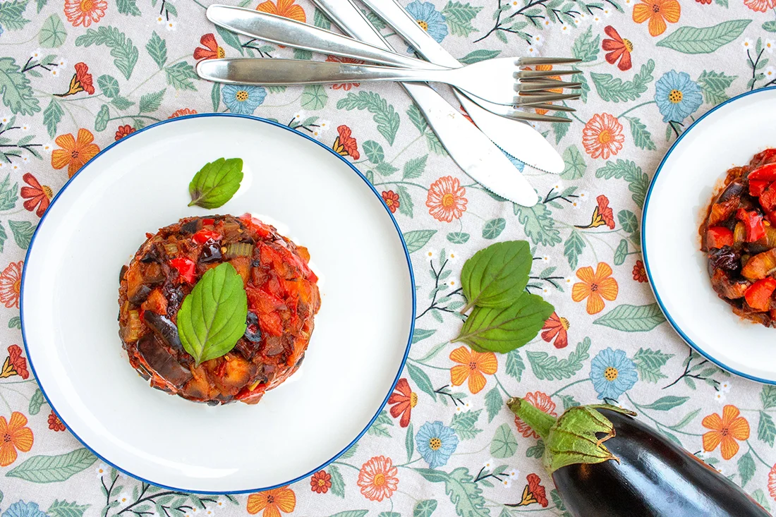 Caponata mit Basilikumblatt auf einem Teller, daneben eine Aubergine und Besteck