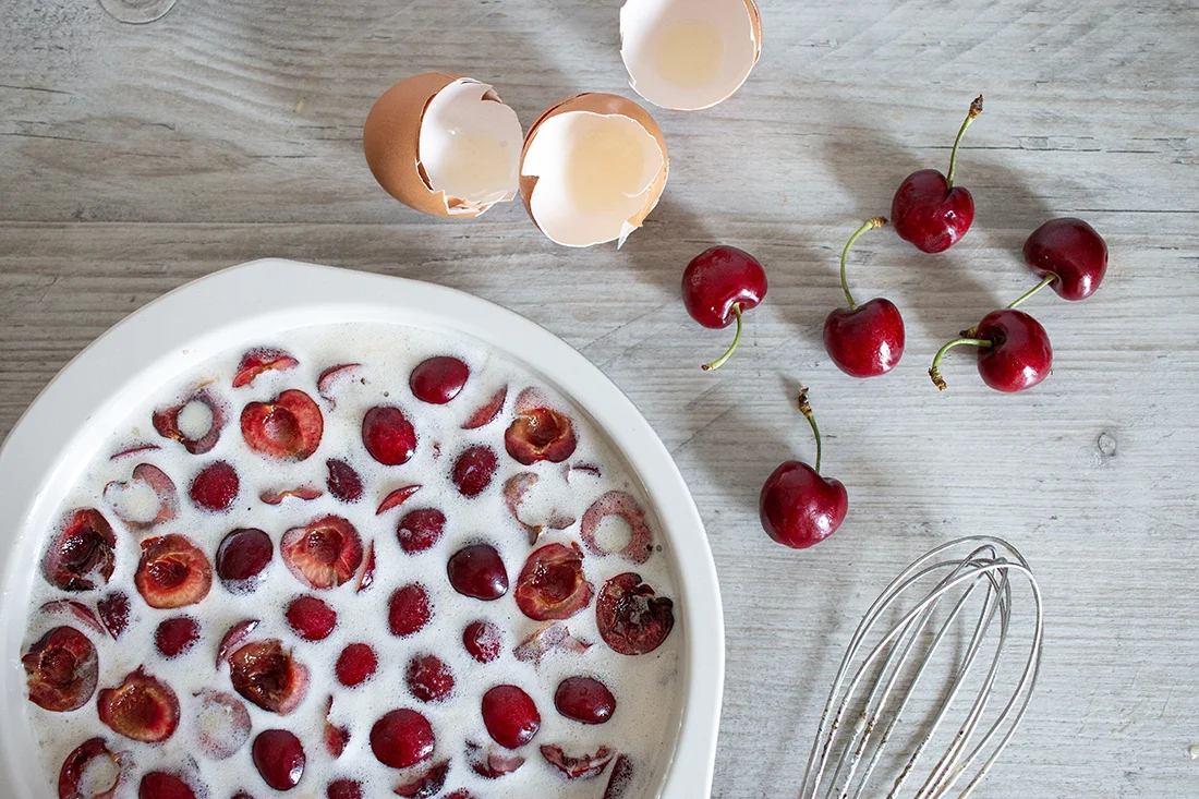 Ungebackener Clafoutis mit Kirschen in einer Form, daneben Eierschalen, ein Schneebesen und frische Kirschen