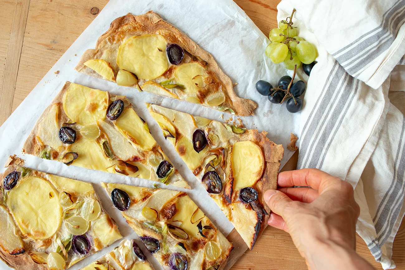 Hand greift ein Stück Flammkuchen mit Kartoffel und Traube von einem geschnittenen Flammkuchen