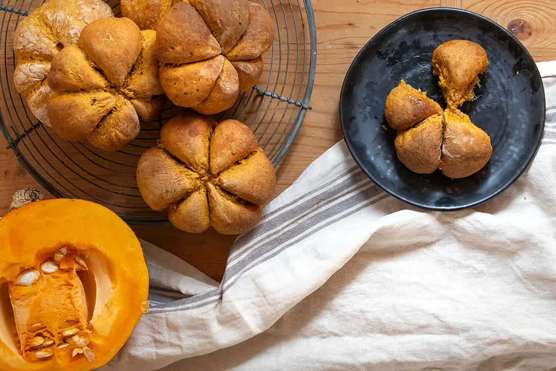 Kürbisbrötchen in Teilen auf einem Teller. Daneben weitere Brötchen und ein halber Kürbis