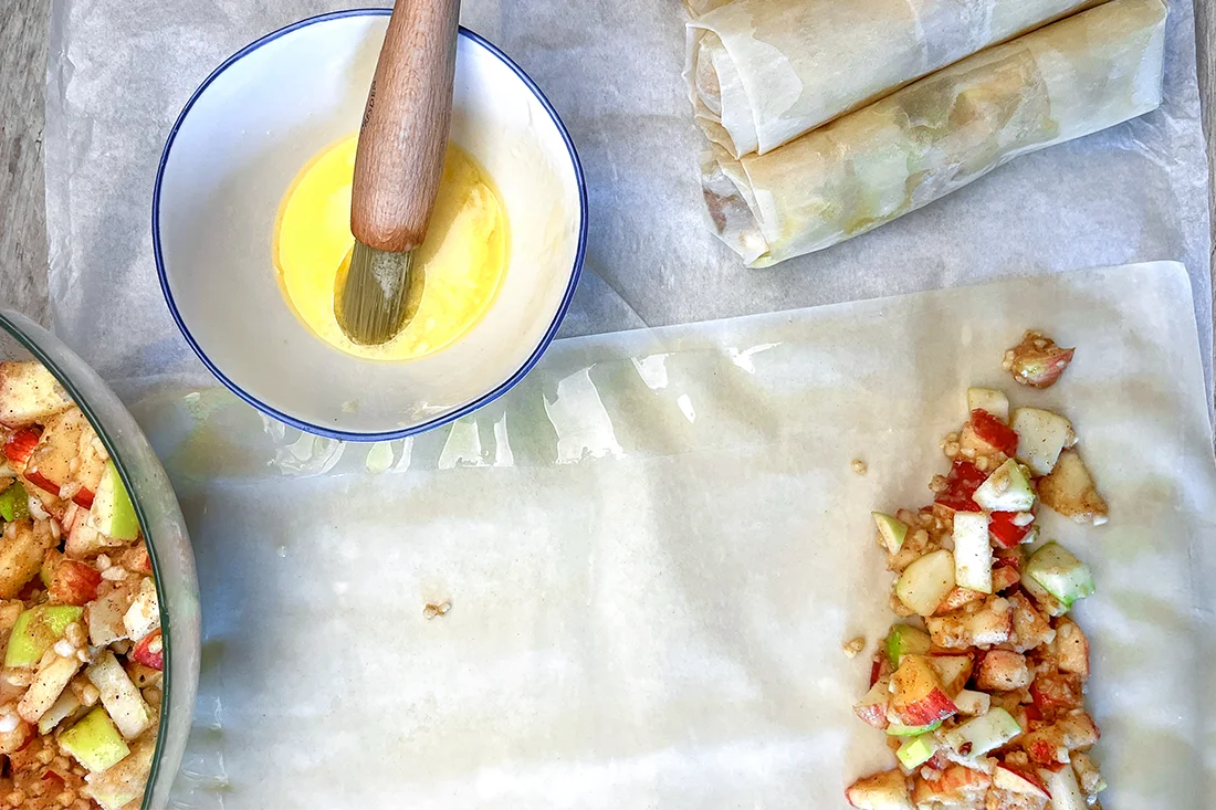 Apfelfüllung auf einer Strudelteigblatte, daneben flüssige Butter in einer Schüssel