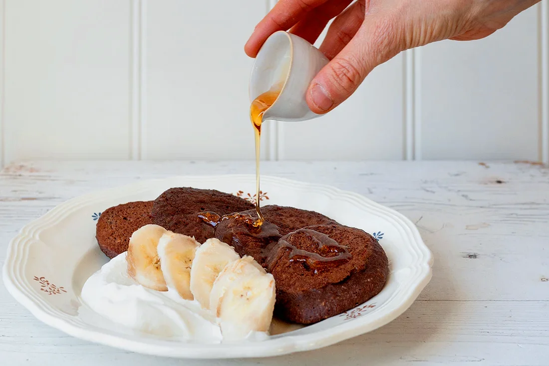 Schoko-Pancakes, Bananenscheiben und vegane Creme, über die etwas Sirup gegossen wird.