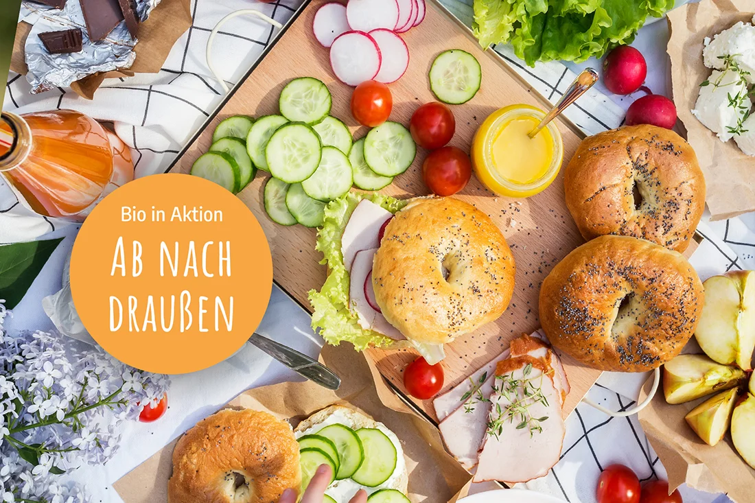 Bagels, Gurkenscheiben, Tomaten und Belag auf einer Picknickdecke in der Draufsicht. Weißer Text auf orangem Kreis. Bio in Aktion: Ab nach draußen. Bild: Adobe-Stock/Irina Schmidt
