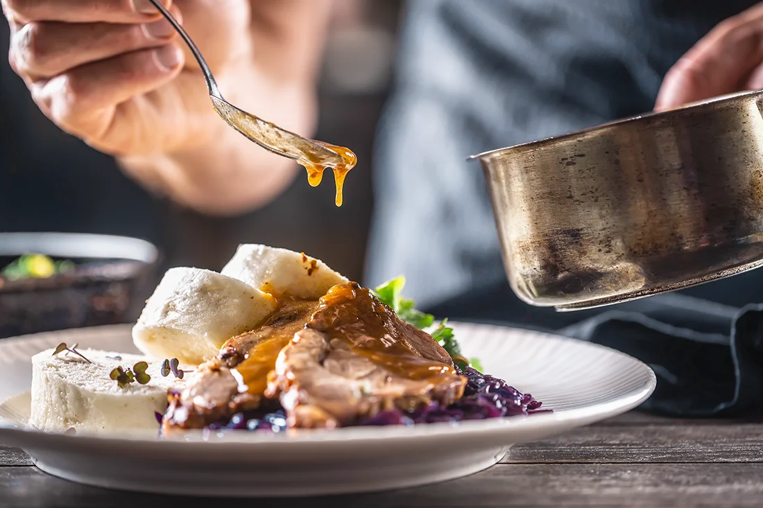 Eine Hand gibt Sauce auf einen Teller mit Knödeln, Rotkohl, Kräutern und Fleisch.