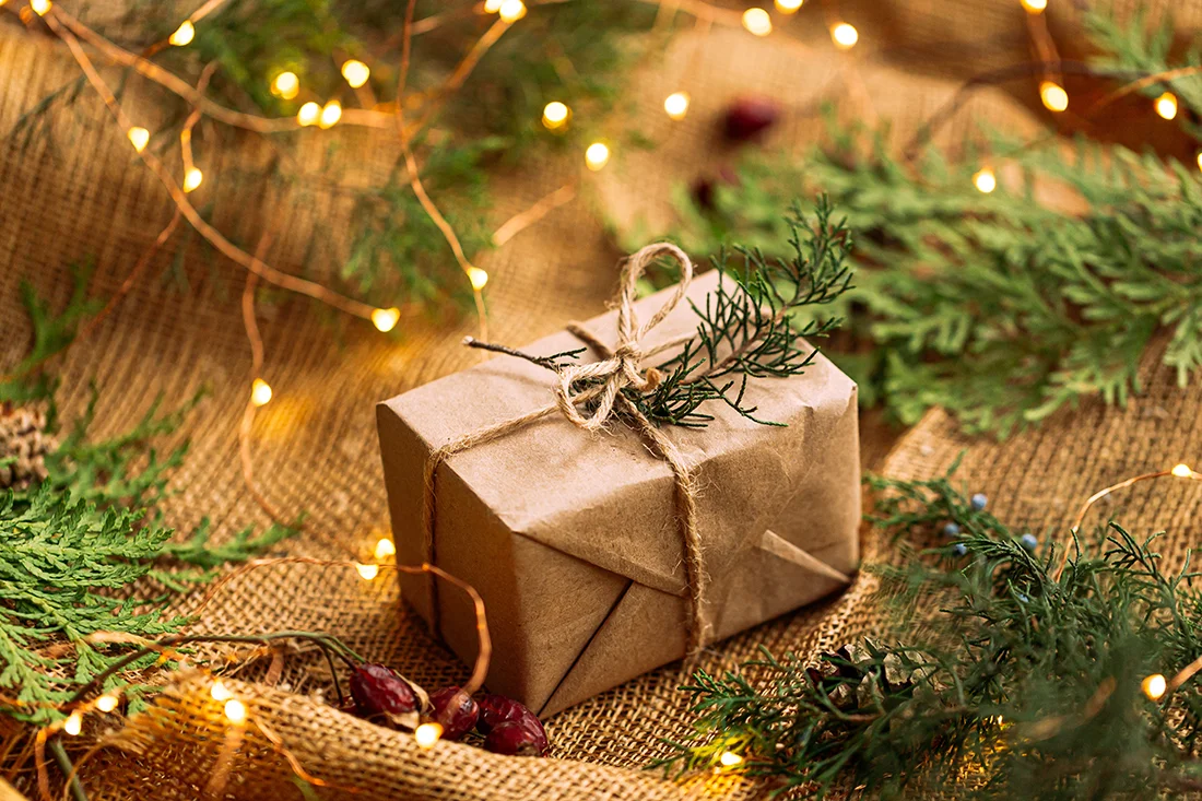 Ein verpacktes Geschenk auf braunem Stoff mit Tannengrün und Lichtern. Bild: AdobeStock_Hihitetlin