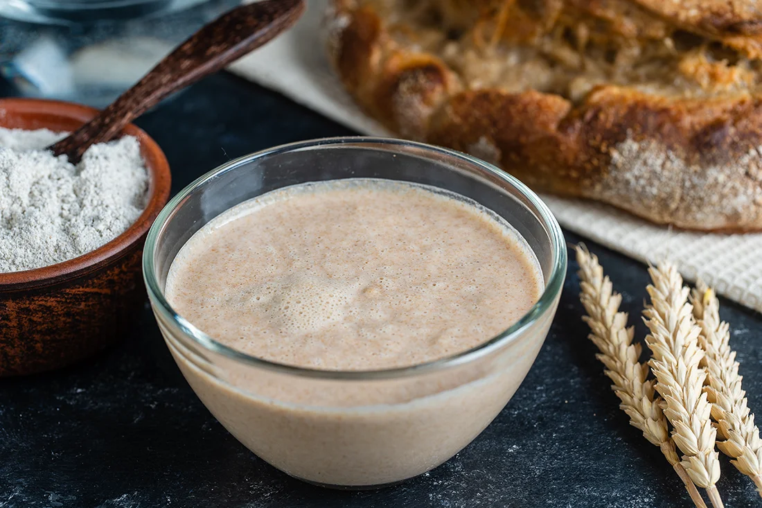 Sauerteig in einer kleinen Schüssel, daneben Mehl und Getreide
