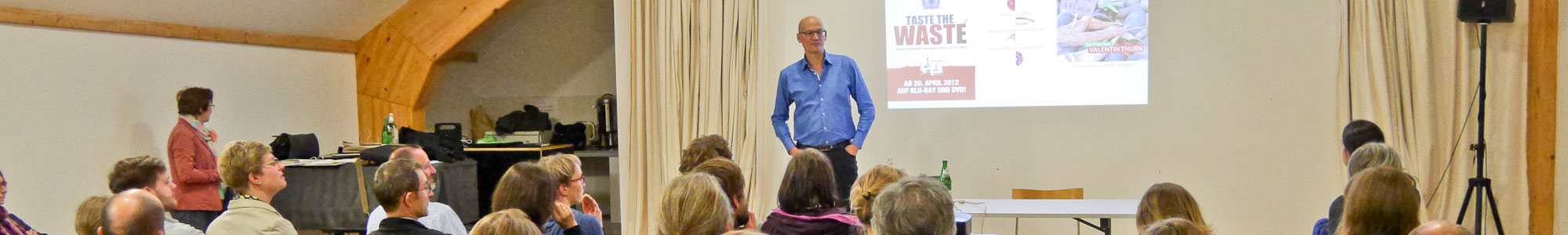 Mann hält einen Vortrag vor sitzenden Menschen. Auf einer Leinwand der Titel des Vortrages "Taste the Waste"